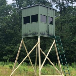 Raised hunting stand with a green cabin atop wooden legs in a grassy field.