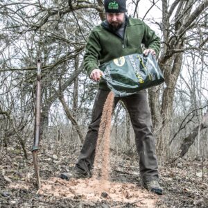 Bearded man in a green fleece jacket pours soil from a bag into a trench in a leaf-strewn yard surrounded by bare trees.