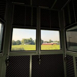 Interior of a soundproof recording booth with foam panels and three windows showing a grassy field and a red building outside