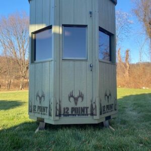 Octagonal green hunting blind on stilts in a grassy field, with three windows and a door, and '12 POINT' branding along the base.