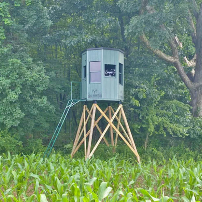 6 octagon - Ideal Land Management | Forestry mowing & mulching, food plots, hunting blinds Elevated green hunting stand on wooden stilts overlooking a cornfield, with a ladder leaning to the cabin among trees.