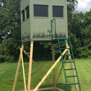 Elevated green hunting blind on a wooden stilt frame with a ladder, overlooking a grassy field.
