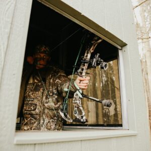 Hunter in camouflage inside a hunting blind, drawing a compound bow toward the woods outside the window