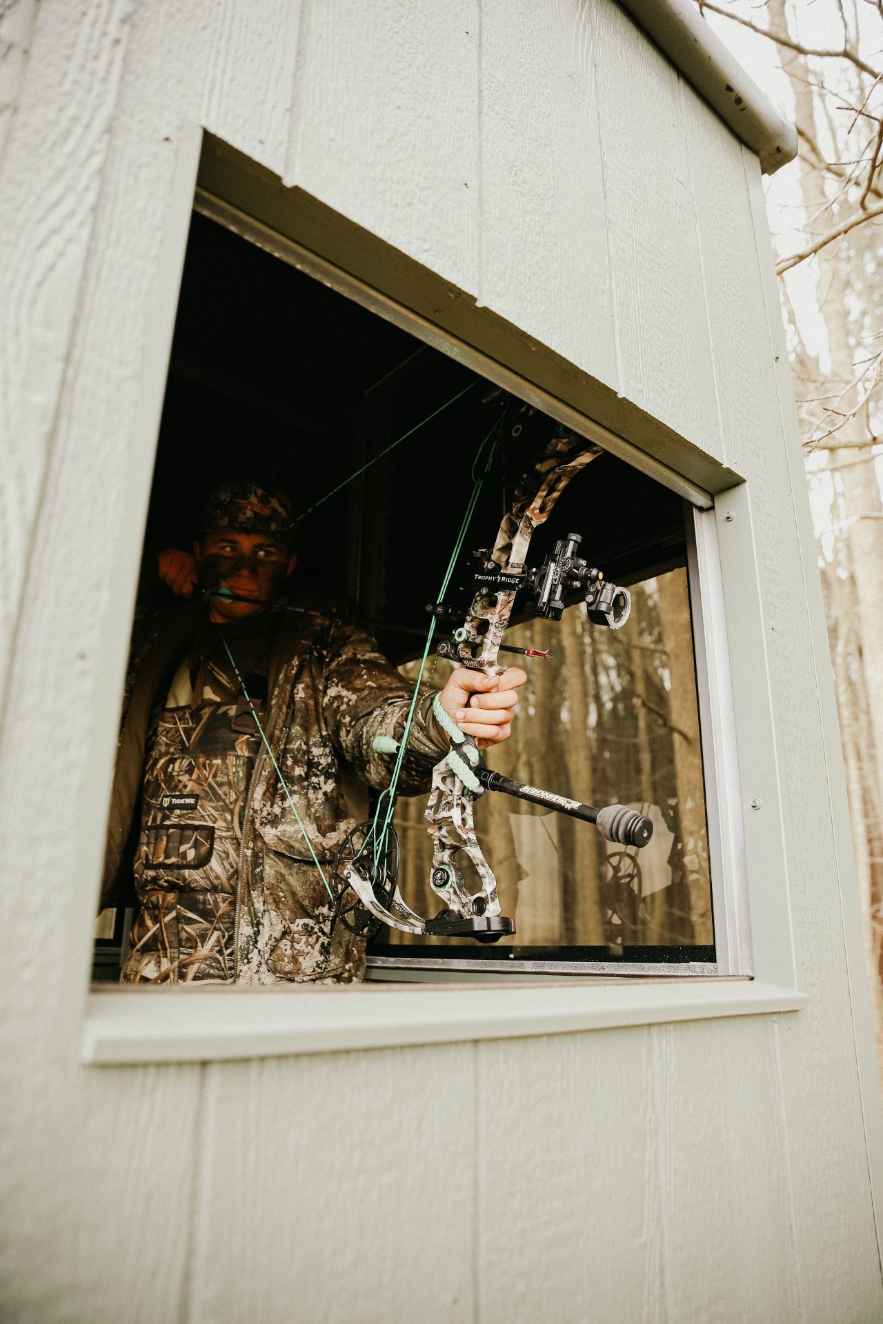 Dallas-Ludwig-IMG_0613 - Ideal Land Management | Forestry mowing & mulching, food plots, hunting blinds Hunter in camouflage inside a hunting blind, drawing a compound bow toward the woods outside the window