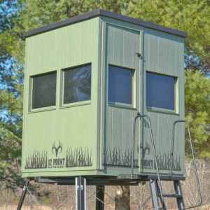 Green elevated hunting blind on a metal stand with windows, set among trees and a ladder for access (12 Point Hunting Blinds).