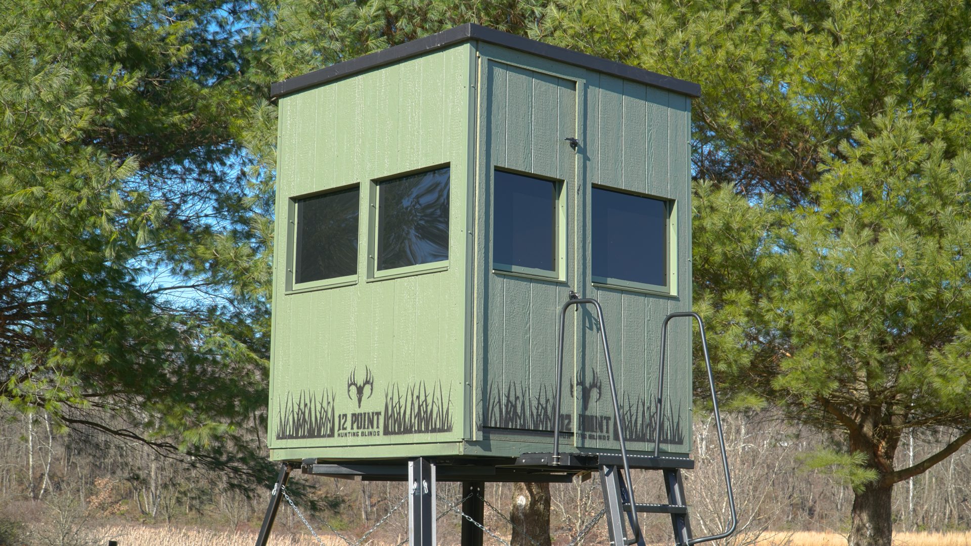 Double-Barrel-N-t-Hero - Ideal Land Management | Forestry mowing & mulching, food plots, hunting blinds Raised green hunting blind on a metal platform with a ladder, set among pine trees.