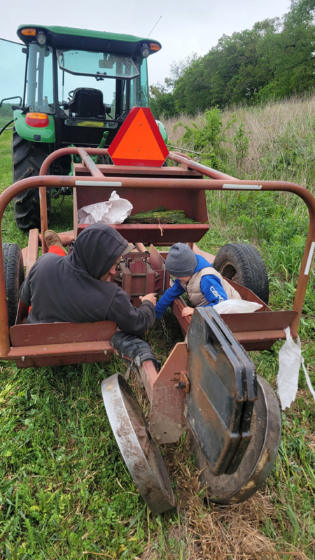 Two people in hooded jackets kneel beside a red farm implement hitched to a green tractor in a grassy field, with an orange warning triangle on top.