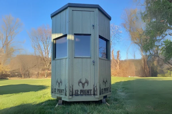 The6_Octagon-12PointHuntingBlinds_1_600x - Ideal Land Management | Forestry mowing & mulching, food plots, hunting blinds Small gray wooden observation tower on a grassy lawn, with three windows and '12 POINT' logos at the base, in a park.