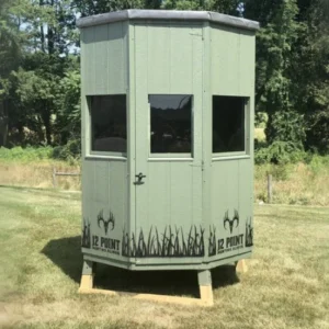 Elevated hunting observation shack on stilts in a grassy field, light green with three windows and deer-antler graphics near the base.