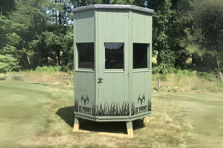 The_6_Octagon_3_1200x - Ideal Land Management | Forestry mowing & mulching, food plots, hunting blinds Elevated hunting observation shack on stilts in a grassy field, light green with three windows and deer-antler graphics near the base.