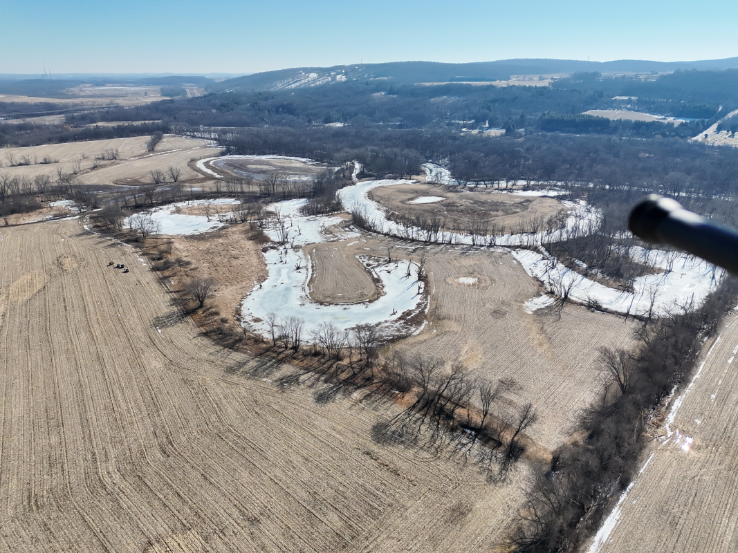 Aerial view of a winding frozen river through bare trees and brown fields with distant hills on the horizon.