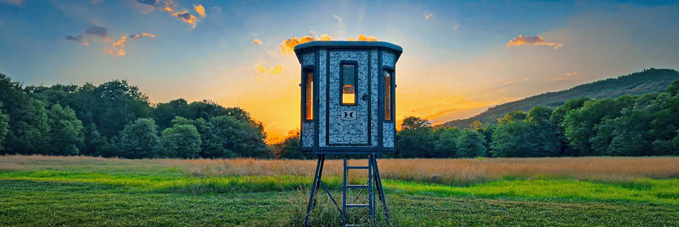 Elevated hunting blind on metal legs in a grassy field at sunset, with a tree line and hills in the background.