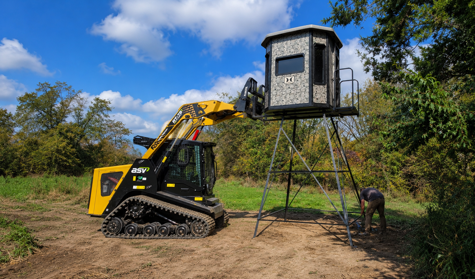 Compact tracked loader with a hydraulic arm lifting a camouflaged hunting blind onto a tall metal stand in a dirt clearing, a person nearby working on the ground.