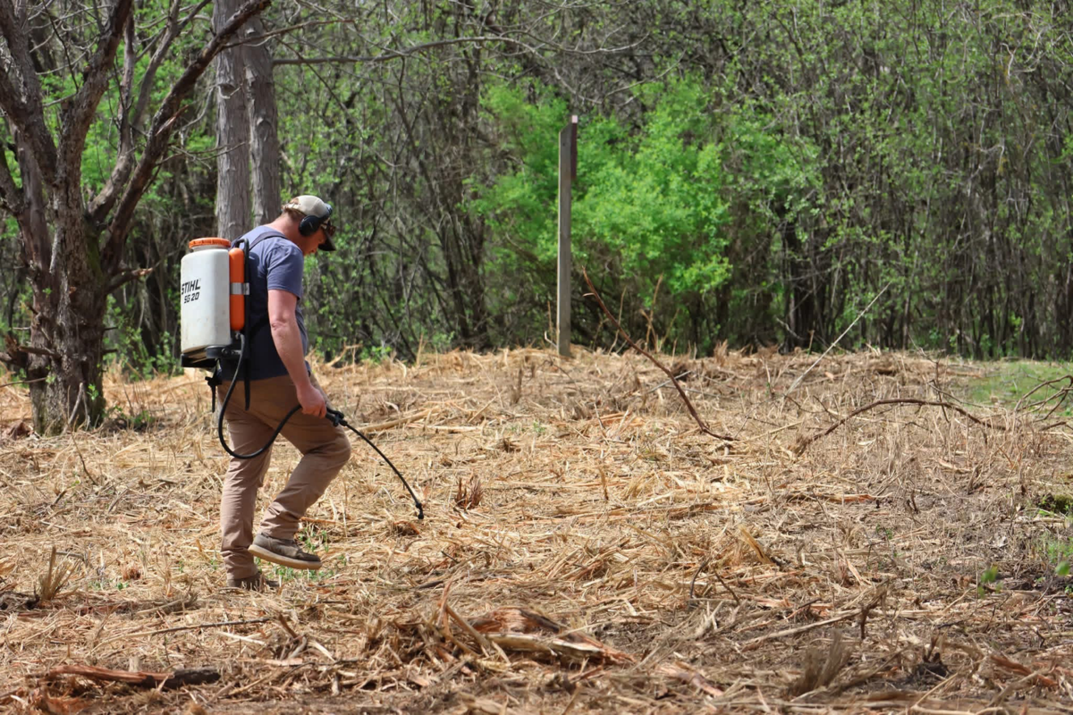A man wearing a cap and headphones walks through a dried grassy field with a backpack sprayer for applying chemicals.