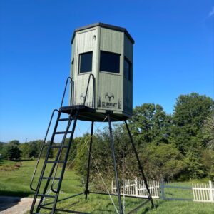 Green elevated hunting blind on metal stilts with a ladder, set in a sunny yard.