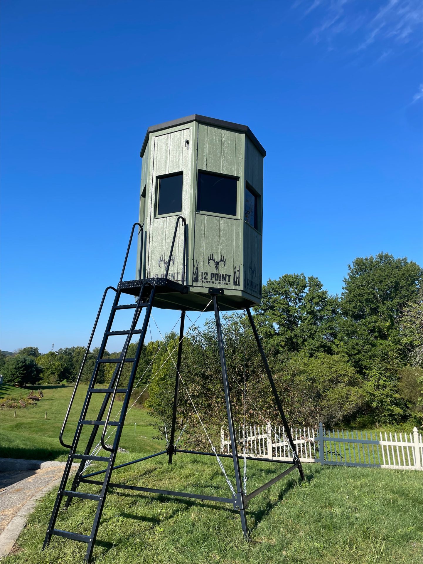 metal-stand-ls - Ideal Land Management | Forestry mowing & mulching, food plots, hunting blinds Green elevated hunting blind on metal stilts with a ladder, set in a sunny yard.